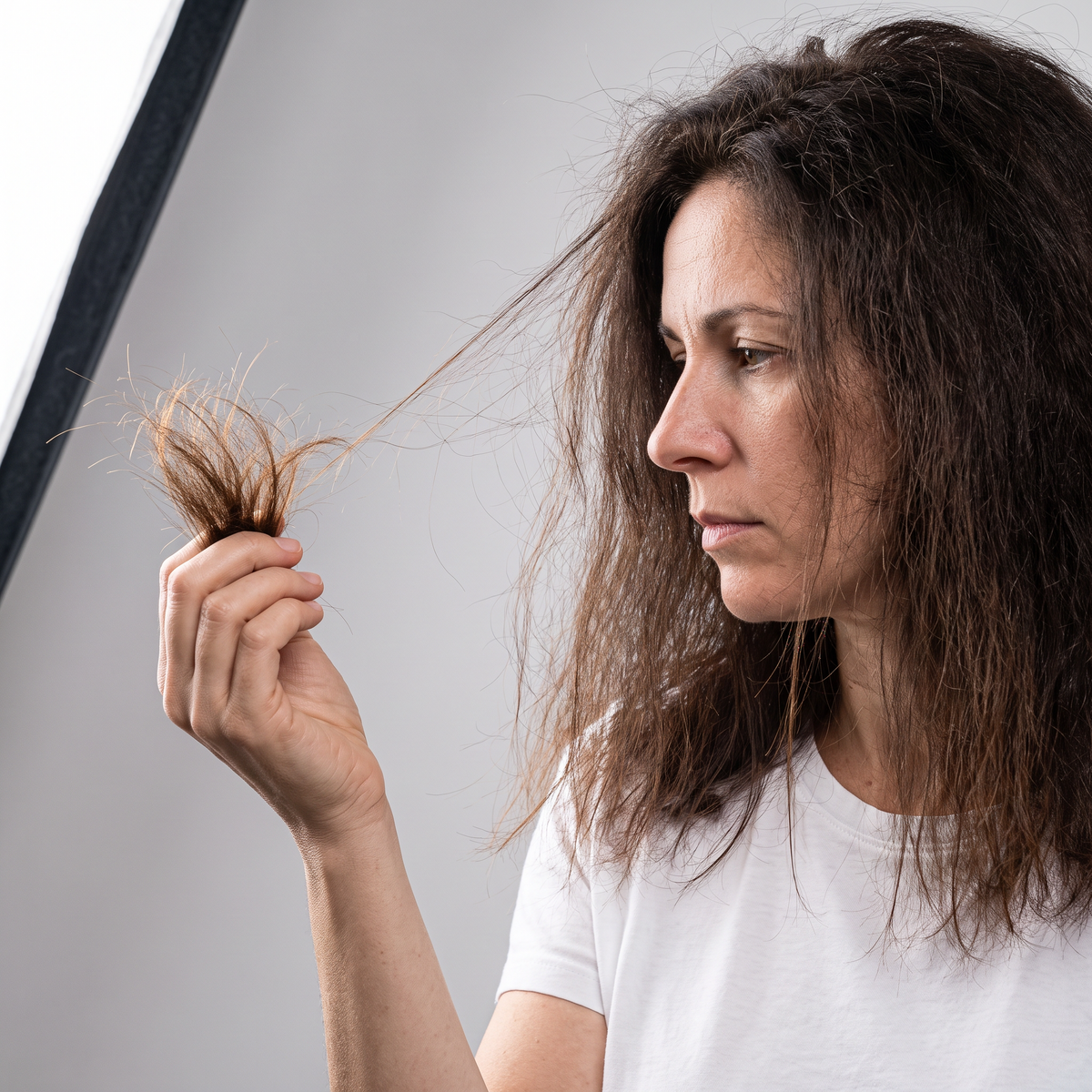 Woman in a white T-shirt holding up a lock of dry, frizzy hair with split ends, before using the Bledna routine